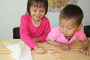 Zwei Kinder sitzen an einem Tisch und schauen auf einen Wüfel, der fünf Augen zeigt