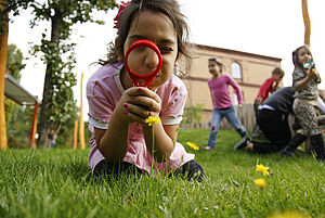 Ein Mädchen betrachtet eine Blume durch eine Lupe. Im Hintergrund laufen mehrere Kinder über die Wiese.
