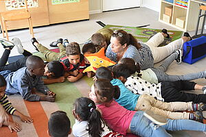 Kinder liegen zusammen mit einer Pädagogin auf dem Boden. Die Pädagogin erzählt etwas und die Kinder hören gespannt zu.