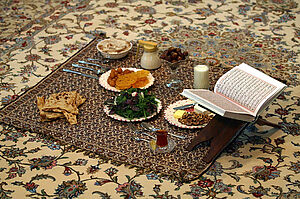 Auf einem Teppich auf dem Boden stehen Datteln, Brot, Salat, Tee und ein Koran